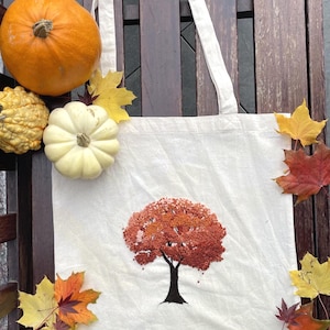 May include: A cream-colored canvas tote bag featuring a central embroidered design of a tree with a dark brown trunk and a dense canopy of orange, red, and rust-colored French knots, surrounded by scattered autumn leaves and two pumpkins.
