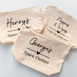 Peut inclure: Trois pochettes zipp&eacute;es en toile beige avec texte noir. L'une porte l'inscription "Henry's Spare", une autre "Nappies & Wipes Things that smell nice", et la troisi&egrave;me "George's Spare Clothes". Chaque pochette a un c&oelig;ur noir.