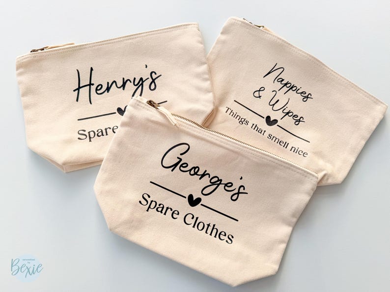 Peut inclure: Trois pochettes zipp&eacute;es en toile beige avec texte noir. L'une porte l'inscription "Henry's Spare", une autre "Nappies & Wipes Things that smell nice", et la troisi&egrave;me "George's Spare Clothes". Chaque pochette a un c&oelig;ur noir.
