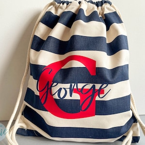 K&ouml;nnte beinhalten: Ein Kordelzugrucksack mit horizontalen marineblauen und cremefarbenen Streifen. Die Tasche ist mit einem gro&szlig;en roten Buchstaben G und dem Namen George in dunkelblauer Schreibschrift in der Mitte personalisiert. Er hat dicke cremefarbene Kordeln.