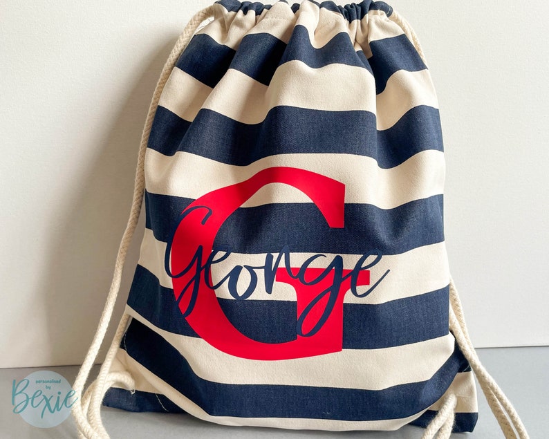 K&ouml;nnte beinhalten: Ein Kordelzugrucksack mit horizontalen marineblauen und cremefarbenen Streifen. Die Tasche ist mit einem gro&szlig;en roten Buchstaben G und dem Namen George in dunkelblauer Schreibschrift in der Mitte personalisiert. Er hat dicke cremefarbene Kordeln.