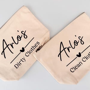 Peut inclure: Deux pochettes zipp&eacute;es en toile beige. L'une porte l'inscription "Arlo's Dirty Clothes" et l'autre "Arlo's Clean Clothes" en &eacute;criture noire. Un c&oelig;ur noir et un trait sont sous le nom. Les pochettes sont sur fond blanc.