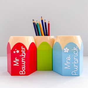 May include: Three pencil-shaped desk tidies in red, green, and blue, each with a cream-coloured top. The red tidy has the text "Mr. Baumber" and stars. The blue tidy has the text "Mrs. Purbrick" and a flower. Pencils are inside.