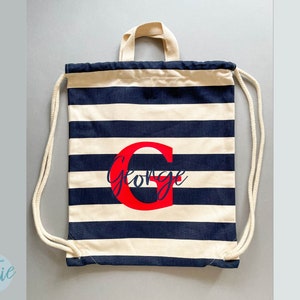 K&ouml;nnte beinhalten: Ein Kordelzugrucksack mit horizontalen marineblauen und cremefarbenen Streifen. Die Tasche hat cremefarbene Kordeltr&auml;ger und einen Tragegriff. Mittig auf der Vorderseite befindet sich ein gro&szlig;er roter Buchstabe G mit dem Namen George in blauer Schrift.