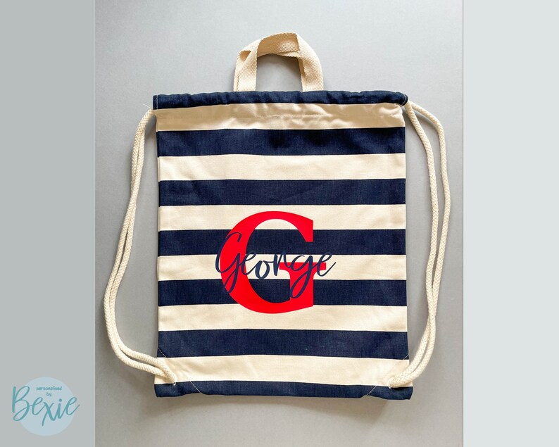 K&ouml;nnte beinhalten: Ein Kordelzugrucksack mit horizontalen marineblauen und cremefarbenen Streifen. Die Tasche hat cremefarbene Kordeltr&auml;ger und einen Tragegriff. Mittig auf der Vorderseite befindet sich ein gro&szlig;er roter Buchstabe G mit dem Namen George in blauer Schrift.