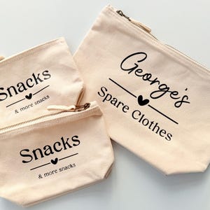 Peut inclure: Trois pochettes zipp&eacute;es en toile beige. Deux petites pochettes indiquent "Snacks & more snacks" en &eacute;criture noire. La plus grande pochette indique "George's Spare Clothes" en &eacute;criture noire. Toutes les pochettes ont un c&oelig;ur noir.