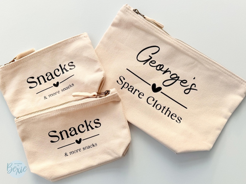 Peut inclure: Trois pochettes zipp&eacute;es en toile beige. Deux petites pochettes indiquent "Snacks & more snacks" en &eacute;criture noire. La plus grande pochette indique "George's Spare Clothes" en &eacute;criture noire. Toutes les pochettes ont un c&oelig;ur noir.