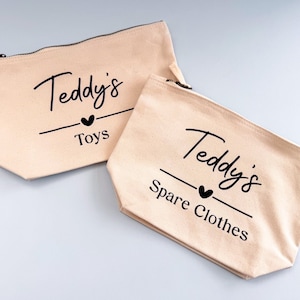 Peut inclure: Deux pochettes zipp&eacute;es en toile beige avec l'inscription noire "Teddy's Toys" et "Teddy's Spare Clothes". Chaque pochette comporte un symbole de c&oelig;ur noir. Les pochettes sont pos&eacute;es sur une surface gris clair.
