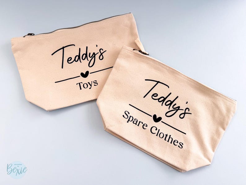 Peut inclure: Deux pochettes zipp&eacute;es en toile beige avec l'inscription noire "Teddy's Toys" et "Teddy's Spare Clothes". Chaque pochette comporte un symbole de c&oelig;ur noir. Les pochettes sont pos&eacute;es sur une surface gris clair.