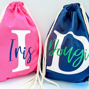 May include: A bright pink drawstring bag with a large white letter I and the name Iris in dark blue script, and a navy blue drawstring bag with a large white letter D and the name Dougie in green script, both featuring white rope straps.