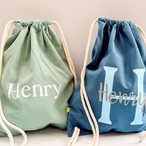 May include: Two drawstring backpacks with thick cream-coloured rope straps. The left bag is sage green with the name Henry printed in white. The right bag is dark blue with a large light blue letter H and the name Henry printed in grey script.