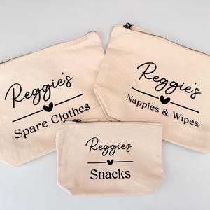 Peut inclure: Trois pochettes zipp&eacute;es en toile beige. Elles portent l'inscription noire "Reggie's", un c&oelig;ur et une ligne, avec les mots "Spare Clothes", "Nappies & Wipes" et "Snacks" en dessous. Diff&eacute;rentes tailles.