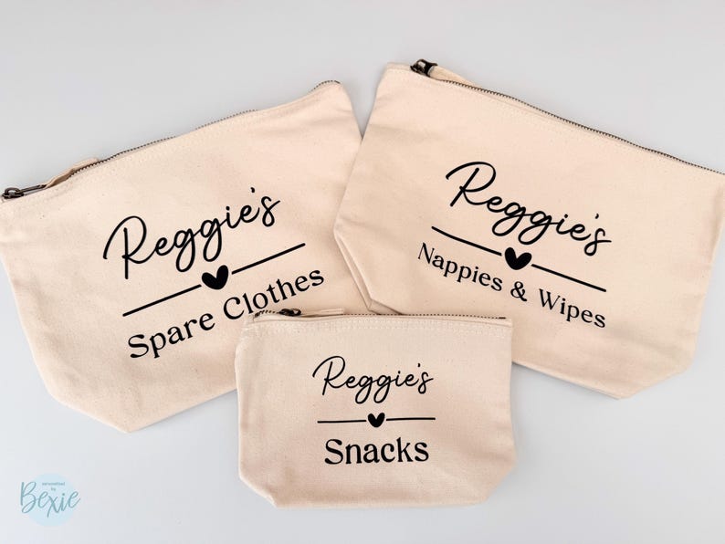 Peut inclure: Trois pochettes zipp&eacute;es en toile beige. Elles portent l'inscription noire "Reggie's", un c&oelig;ur et une ligne, avec les mots "Spare Clothes", "Nappies & Wipes" et "Snacks" en dessous. Diff&eacute;rentes tailles.