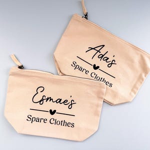 Peut inclure: Deux pochettes zipp&eacute;es en toile beige. L'une porte l'inscription "Esmae's Spare Clothes" en &eacute;criture noire avec un c&oelig;ur. L'autre porte l'inscription "Ada's Spare Clothes" en &eacute;criture noire avec un c&oelig;ur. Les deux ont une tirette beige.