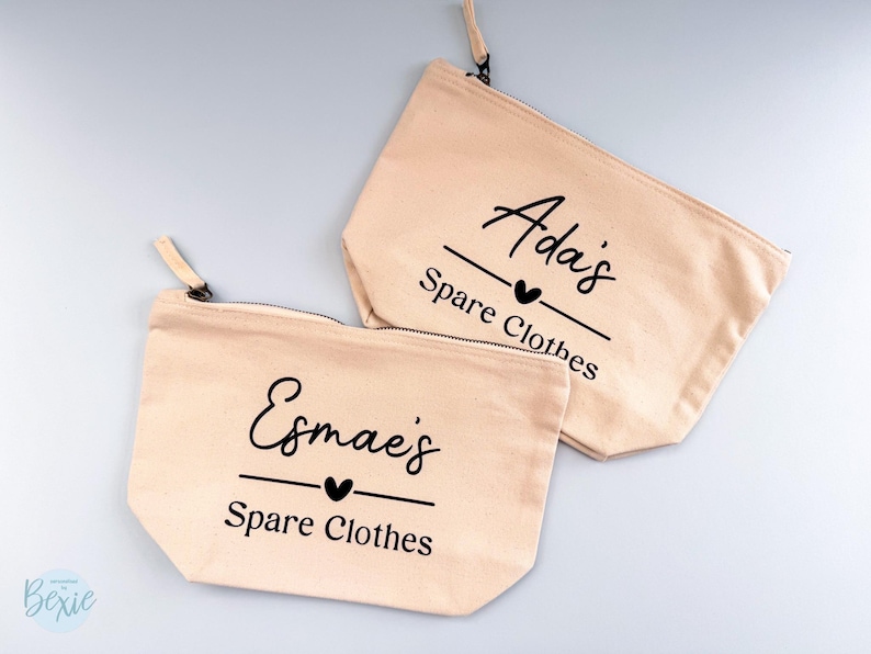 Peut inclure: Deux pochettes zipp&eacute;es en toile beige. L'une porte l'inscription "Esmae's Spare Clothes" en &eacute;criture noire avec un c&oelig;ur. L'autre porte l'inscription "Ada's Spare Clothes" en &eacute;criture noire avec un c&oelig;ur. Les deux ont une tirette beige.