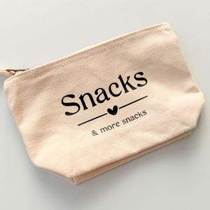 Peut inclure: Une pochette zipp&eacute;e en toile beige avec le mot "Snacks" imprim&eacute; en noir, soulign&eacute; d'un c&oelig;ur, et la phrase "& more snacks" en dessous. La tirette de la fermeture &eacute;clair est beige.
