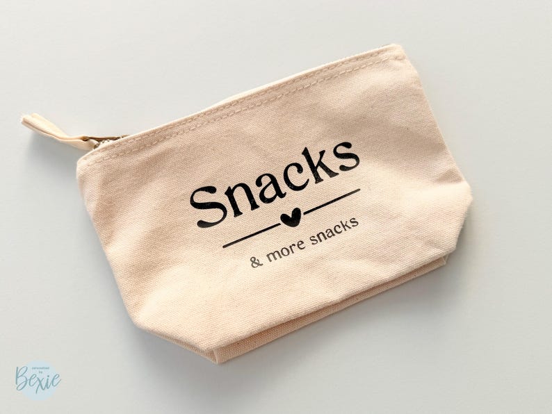 Peut inclure: Une pochette zipp&eacute;e en toile beige avec le mot "Snacks" imprim&eacute; en noir, soulign&eacute; d'un c&oelig;ur, et la phrase "& more snacks" en dessous. La tirette de la fermeture &eacute;clair est beige.