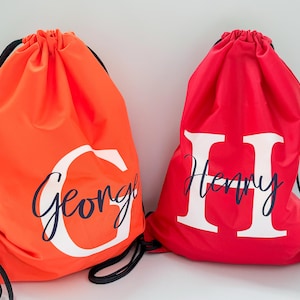 May include: Two drawstring bags are shown, one orange and one red. The orange bag features the name "George" in white cursive with a large white "G". The red bag displays the name "Henry" in white cursive with a large white "H".