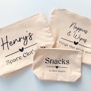 Peut inclure: Trois pochettes zipp&eacute;es en toile beige avec texte noir. La plus grande dit "Nappies & Wipes" et "Things that smell nice". La pochette moyenne dit "Henry's Spare Clot'" et la plus petite dit "Snacks & more snacks."