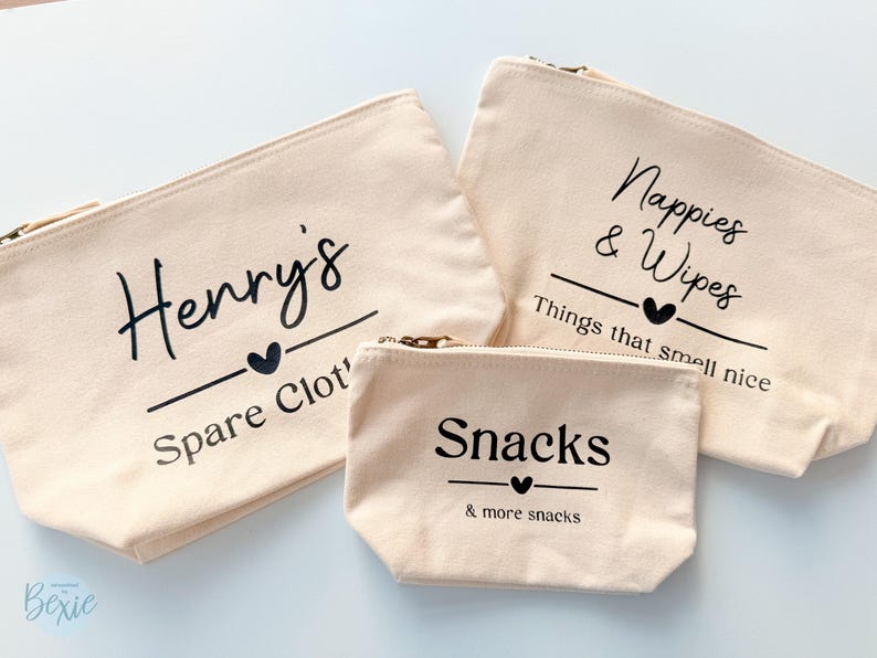 Peut inclure: Trois pochettes zipp&eacute;es en toile beige avec texte noir. La plus grande dit "Nappies & Wipes" et "Things that smell nice". La pochette moyenne dit "Henry's Spare Clot'" et la plus petite dit "Snacks & more snacks."
