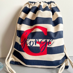 K&ouml;nnte beinhalten: Ein Kordelzugrucksack mit horizontalen marineblauen und cremefarbenen Streifen. Die Tasche ist mit einem gro&szlig;en roten Gro&szlig;buchstaben G und dem Namen George in einer dunkelblauen Schreibschrift in der Mitte personalisiert. Er hat dicke cremefarbene Kordeln.