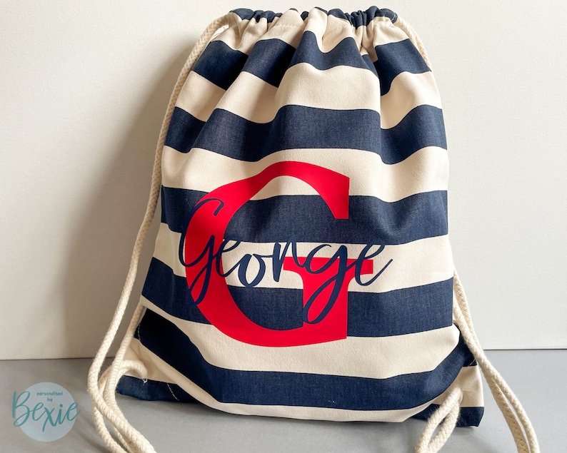 K&ouml;nnte beinhalten: Ein Kordelzugrucksack mit horizontalen marineblauen und cremefarbenen Streifen. Die Tasche ist mit einem gro&szlig;en roten Gro&szlig;buchstaben G und dem Namen George in einer dunkelblauen Schreibschrift in der Mitte personalisiert. Er hat dicke cremefarbene Kordeln.