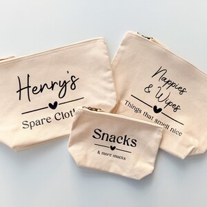 Peut inclure: Trois pochettes zipp&eacute;es en toile beige avec texte noir. La plus grande dit "Nappies & Wipes", celle du milieu dit "Henry's Spare Clot", et la plus petite dit "Snacks & more snacks". Chacune a un symbole de c&oelig;ur.