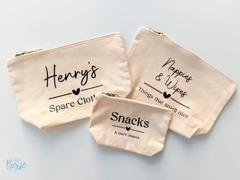 Peut inclure: Trois pochettes zipp&eacute;es en toile beige avec texte noir. La plus grande dit "Nappies & Wipes", celle du milieu dit "Henry's Spare Clot", et la plus petite dit "Snacks & more snacks". Chacune a un symbole de c&oelig;ur.