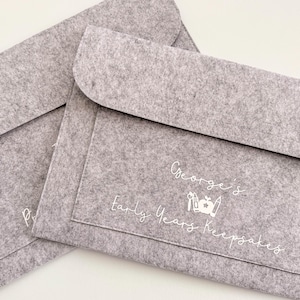 May include: Two grey felt document holders with a flap closure. One holder has the name "George's Early Years Keepsakes" printed in white script. The other holder has the letter "P" printed in white script.