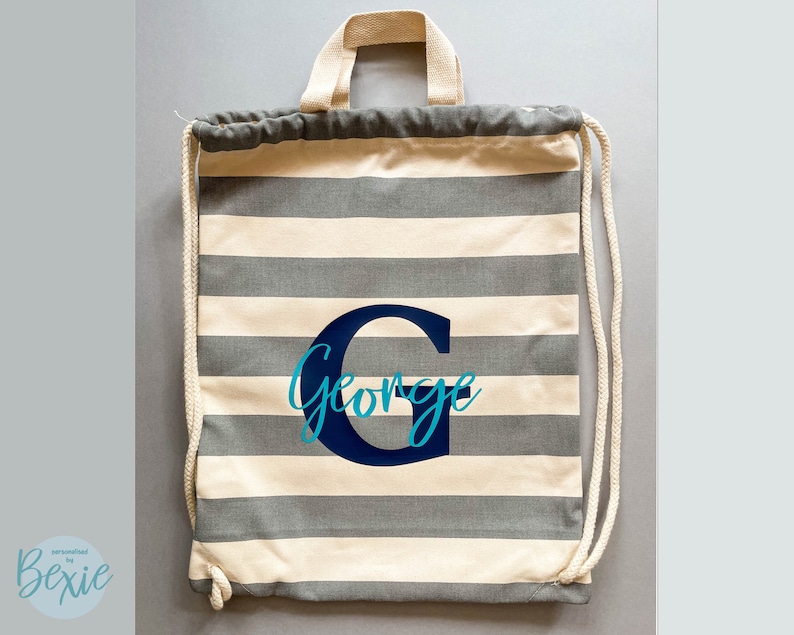 K&ouml;nnte beinhalten: Ein Kordelzugrucksack mit horizontalen grauen und cremefarbenen Streifen. Die Tasche hat cremefarbene gewebte Griffe und passende Kordelz&uuml;ge. In der Mitte befindet sich ein gro&szlig;er dunkelblauer Buchstabe G mit dem Namen George in T&uuml;rkis.