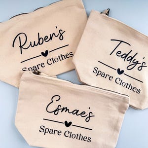 Peut inclure: Trois pochettes zipp&eacute;es en toile beige avec texte noir. Chaque pochette porte un nom, un c&oelig;ur et les mots "Spare Clothes". Les noms sont Ruben's, Teddy's et Esmae's. Les pochettes sont sur une surface bleu clair.