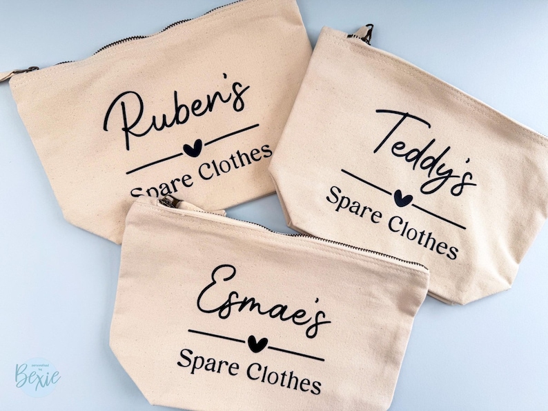 Peut inclure: Trois pochettes zipp&eacute;es en toile beige avec texte noir. Chaque pochette porte un nom, un c&oelig;ur et les mots "Spare Clothes". Les noms sont Ruben's, Teddy's et Esmae's. Les pochettes sont sur une surface bleu clair.