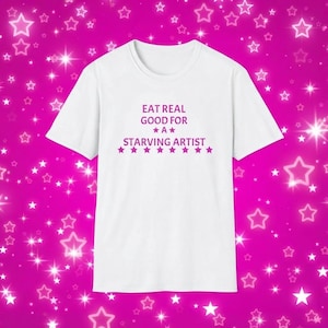 May include: A white short-sleeved t-shirt featuring the purple text EAT REAL GOOD FOR A STARVING ARTIST centred on the chest. The design includes a single purple star above and below the letter A, and a row of nine purple stars below the text.