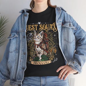 Könnte beinhalten: Schwarzes T-Shirt mit einer detaillierten Illustration einer Katze mit Rucksack und den Worten "QUEST SQUAD" in goldener Schrift. Das Wort "BROCCOLI" befindet sich auf einem Banner unter der Katze. Das Design hat ein Fantasy-Thema.