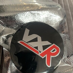 May include: A black round emblem with the letters "VXR" in silver and red. The "V" is on top of the "X" and the "R" is on the bottom. The "R" is red with a silver outline.