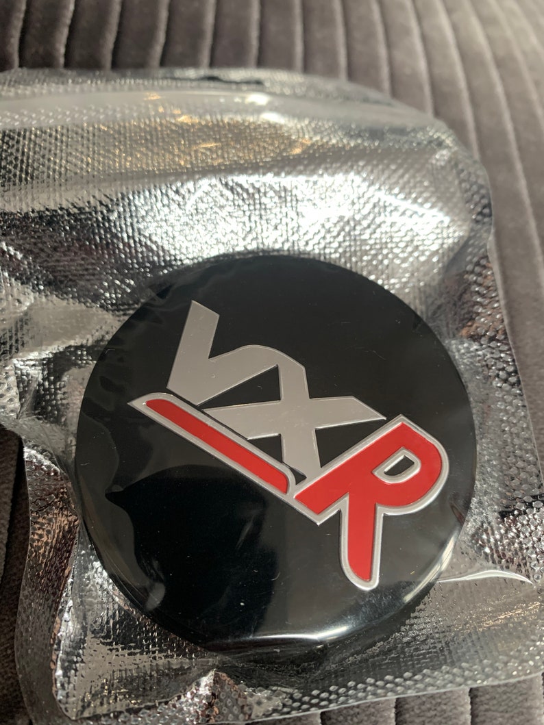 May include: A black round emblem with the letters "VXR" in silver and red. The "V" is on top of the "X" and the "R" is on the bottom. The "R" is red with a silver outline.