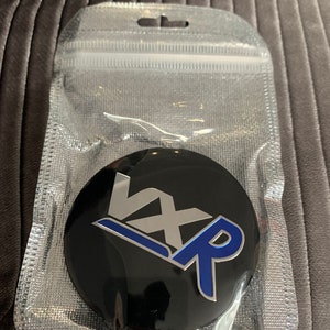 May include: A black round emblem with a silver "VX" and a blue "R" in a stylised design. The emblem is in a clear plastic bag.