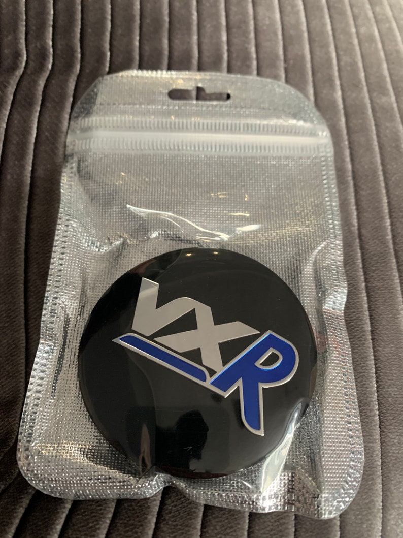 May include: A black round emblem with a silver "VX" and a blue "R" in a stylised design. The emblem is in a clear plastic bag.