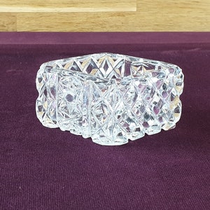 May include: A clear, cut-glass trinket dish with a diamond pattern. The dish has a rectangular shape with a scalloped edge. The glass sparkles, reflecting light. The dish is likely used for jewelry or small items.