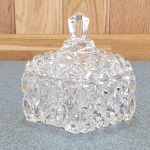 May include: A clear, faceted glass jar with a lid. The jar has a square base and a diamond pattern. The lid has a diamond-shaped handle. The jar is on a grey surface.