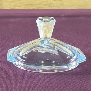 May include: A clear, light blue glass lid with a geometric design. The lid has a faceted, conical handle and a flat, octagonal base. The lid is likely for a decorative container or dish.