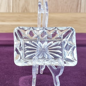 May include: A clear, rectangular glass dish with a starburst design in the centre and diamond-shaped patterns around the edges. The dish is displayed on a clear plastic stand, set against a purple surface and a wooden background.