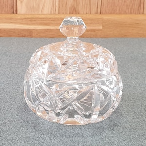 May include: A clear, cut-glass candy dish with a lid. The dish has a round shape with a faceted design, and a knob on the lid. The dish is sitting on a grey surface.