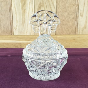 May include: A clear, cut-glass jar with a lid. The jar has a round base and a lid with a decorative handle. The glass is cut with a geometric pattern. The jar is sitting on a purple surface.