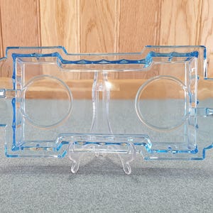 May include: A rectangular, light blue glass serving tray with two circular indentations and two handles. The tray is supported by a clear, plastic stand. The background is a light wood panel.