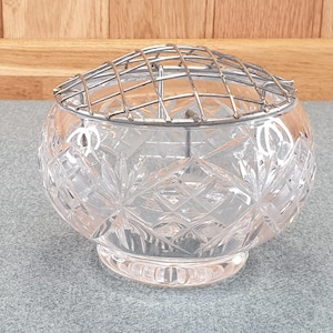 May include: A clear, cut-glass flower frog with a silver metal grid. The round, crystal bowl features a detailed, geometric pattern. The flower arranging tool sits on a grey surface, with a wooden background.