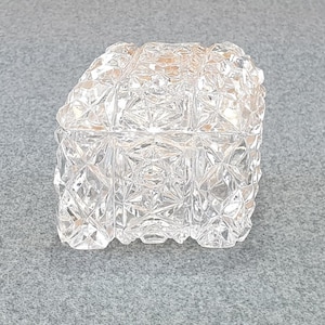 May include: A clear, square crystal trinket box with a textured, geometric pattern. The lid is also textured and fits snugly on the base. The box is displayed on a grey surface.