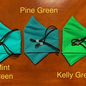 May include: Three face masks in different shades of green. The masks are made of fabric and have adjustable ear loops. The colors are labeled "Mint Green", "Pine Green", and "Kelly Green".