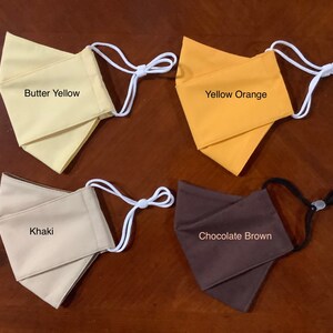 May include: Four fabric face masks in different colors: butter yellow, yellow orange, khaki, and chocolate brown. Each mask has white elastic ear loops.