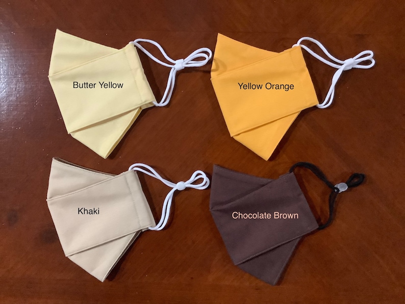 May include: Four fabric face masks in different colors: butter yellow, yellow orange, khaki, and chocolate brown. Each mask has white elastic ear loops.